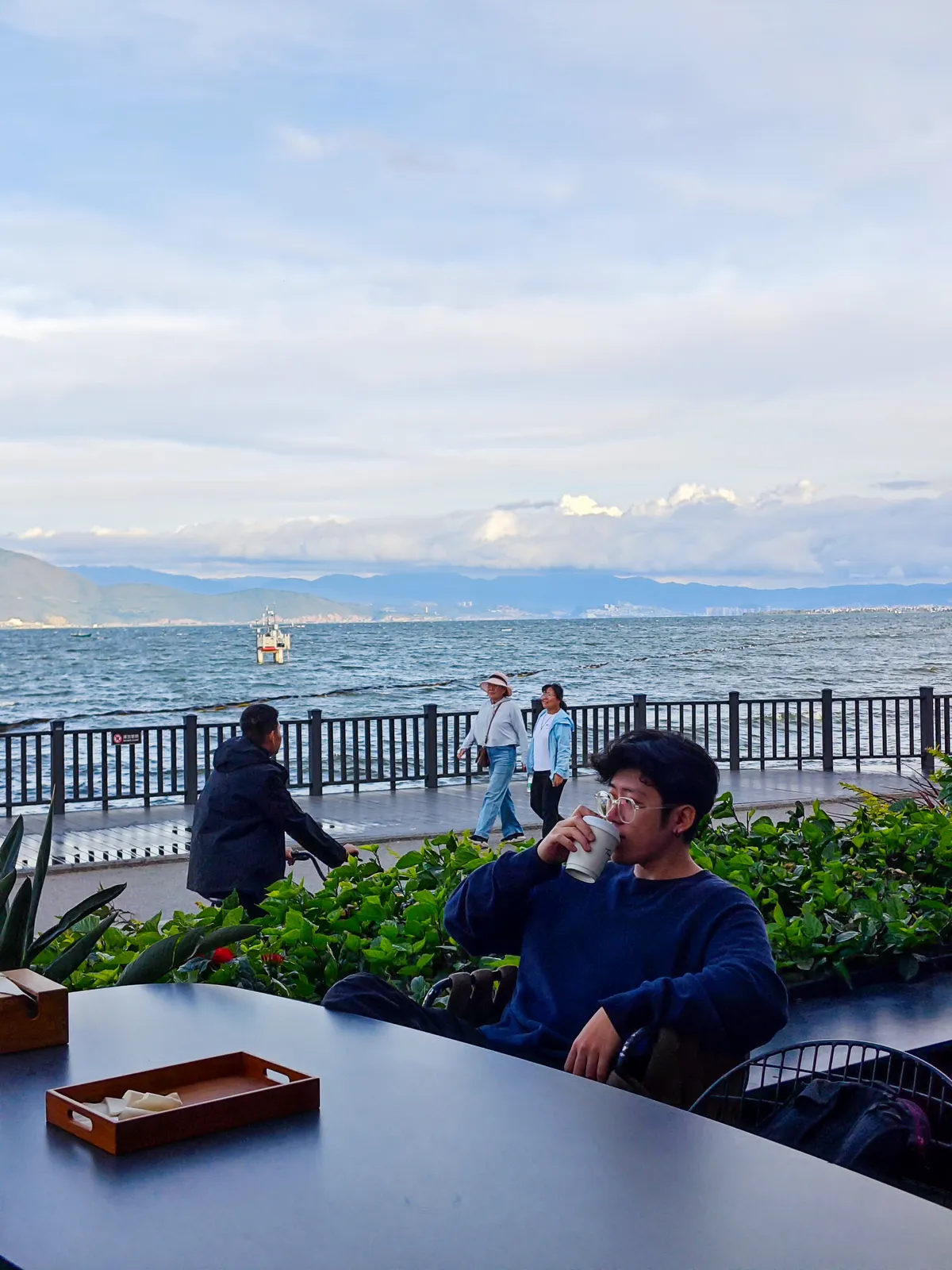 Lin at an outdoor café table by a lake, drinking a coffee, with people walking along the railing behind.
